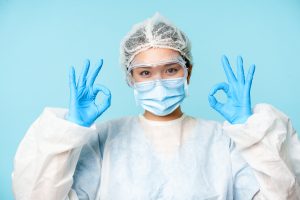 Okay. Smiling asian female doctor, physician in personal protective equipment, showing ok sign, standing over blue background.